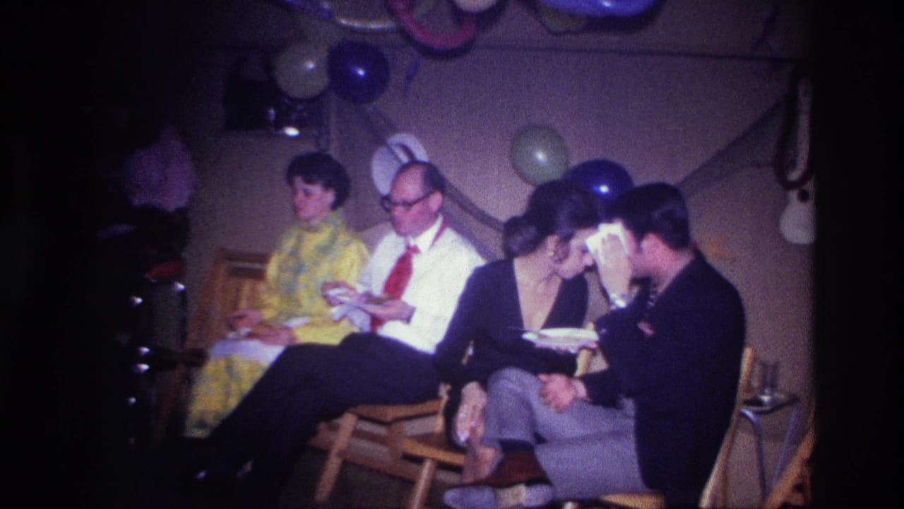 Seated party guests eat and talk beneath balloons and streamers in a dim room.