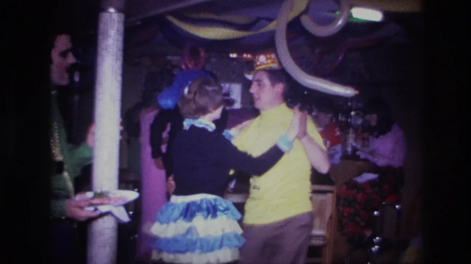 The dancing couple continues in the Ottawa party room, surrounded by balloons, streamers, a column, and seated guests in the background.
