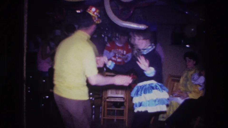 A handheld 8mm frame shows the Ottawa party dancers beneath a long balloon, with guests and stools in the dim background.