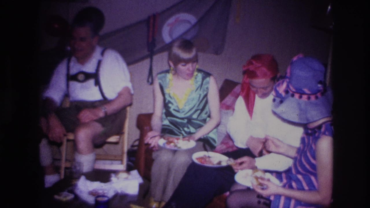 1967 Ottawa home-movie frame showing adults in costume seated indoors, eating from paper plates beside balloons and wall decorations.