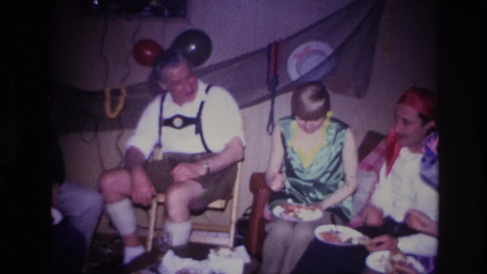 1967 Ottawa home-movie frame showing a man in lederhosen seated beside costumed party guests eating in a private decorated room.
