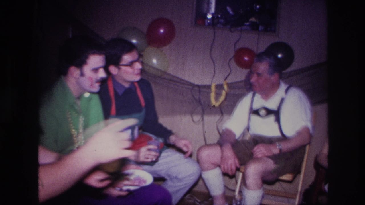 1967 Ottawa home-movie frame showing adult men seated at an indoor party, including a man in lederhosen-style suspenders and balloons behind them.