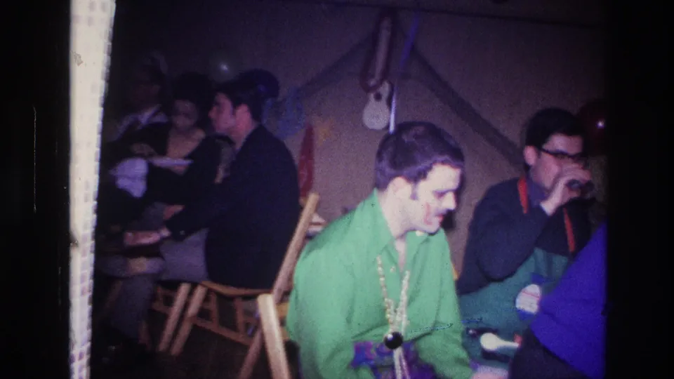 1967 Ottawa home-movie frame showing an indoor party with seated adults, a man in green and beads in front, and guests conversing behind him.