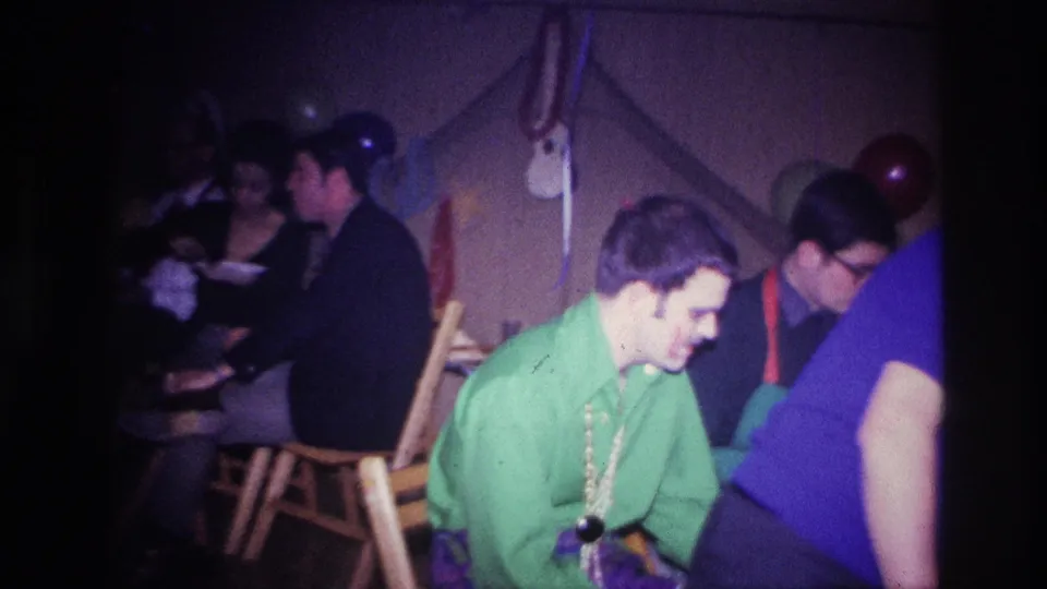 1967 Ottawa party frame with seated adults, a man in green at center, a person in blue partly blocking the view, and balloons on the wall.