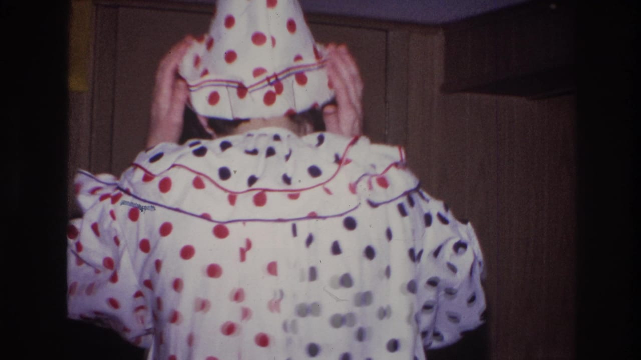 Adult in a white red-and-black polka-dot clown outfit turns away from the camera and adjusts a pointed hat in the Ottawa home-movie party scene.