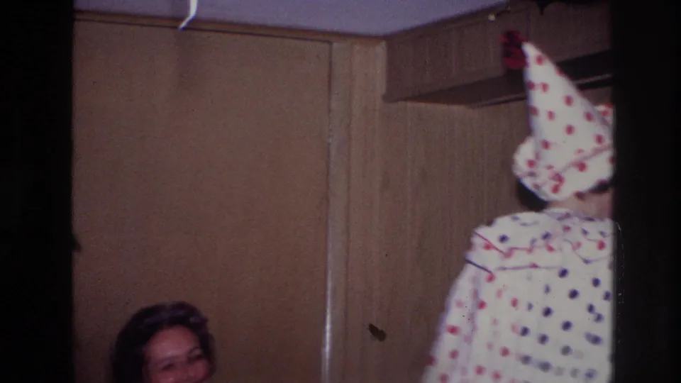 In a wood-paneled private room, the costumed adult is partly out of frame while a seated woman appears at the lower left of the 1967 Ottawa party footage.