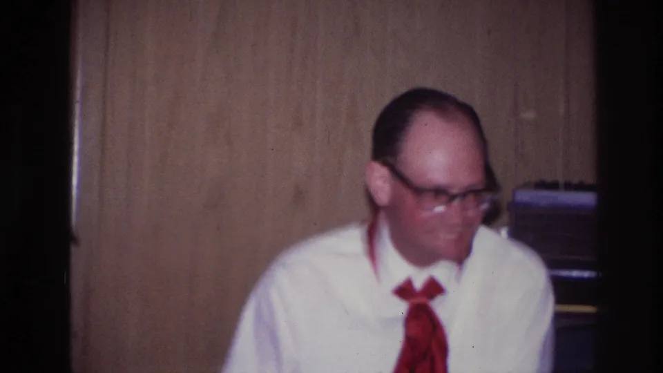 Adult man in glasses, white shirt, and red tie sits against wood paneling during the Ottawa 1967 indoor celebration footage.
