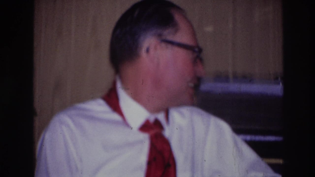 Ottawa 1967 home-movie frame of a seated man wearing glasses, a white shirt, and red neckwear, turned toward someone off camera indoors.