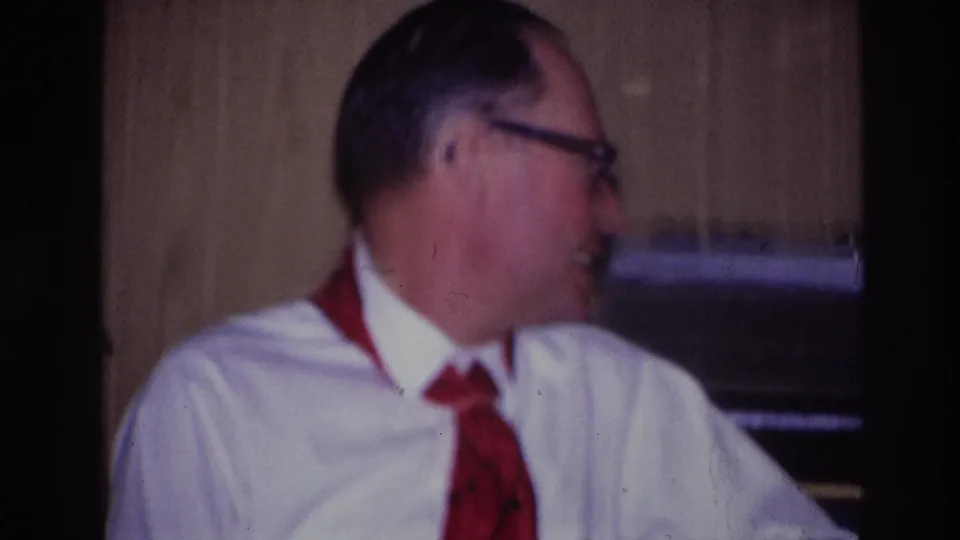 Ottawa 1967 home-movie frame of a seated man wearing glasses, a white shirt, and red neckwear, turned toward someone off camera indoors.