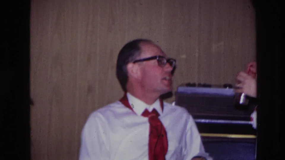 Ottawa 1967 home-movie frame with the seated man looking toward a nearby person whose hand and drink are partly visible.