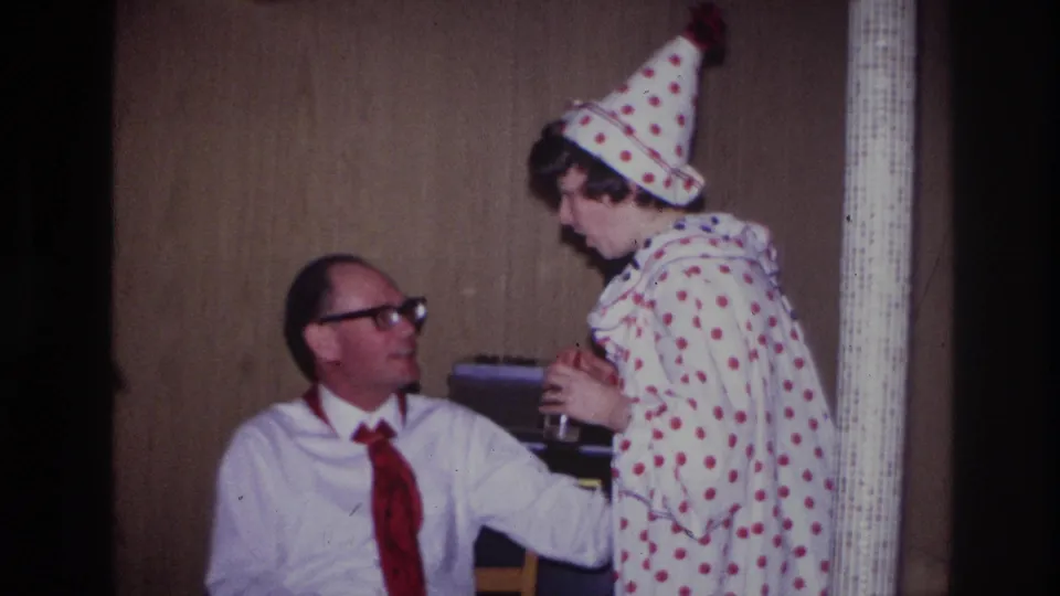 Ottawa 1967 frame showing a costumed young person in a white polka-dot clown outfit and pointed hat standing beside the seated man.