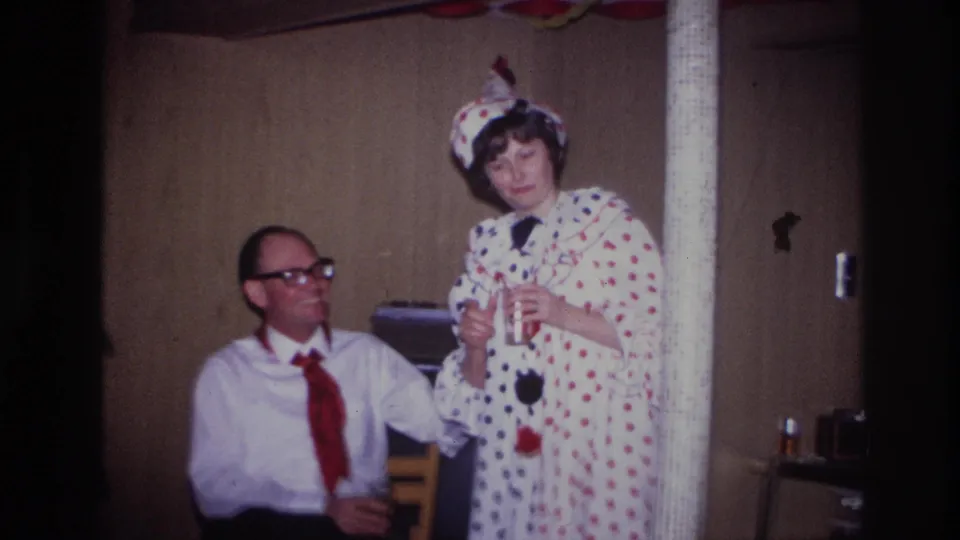 In 1967 Ottawa home-movie footage, a woman in a white polka-dot clown costume stands near a seated man during an indoor party.
