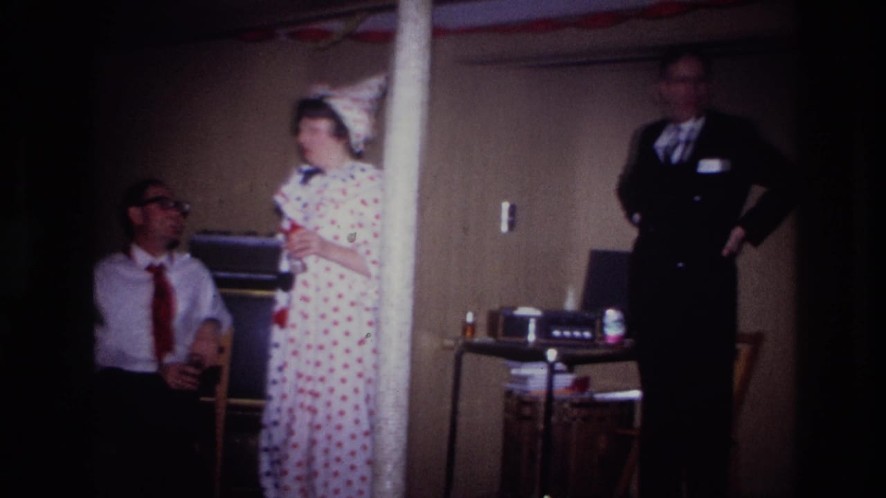 Costumed woman and seated man look toward a man in a dark suit near a small table.