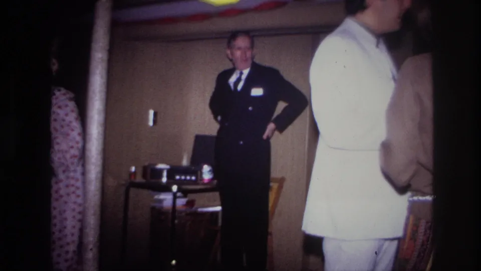 A suited man stands with one hand at his waist while guests in light clothing move across the foreground of the 1967 Ottawa party footage.