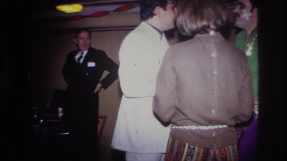 Adults in white, tan, and green clothing gather closely in the foreground while a suited man stands behind them in the Ottawa interior.