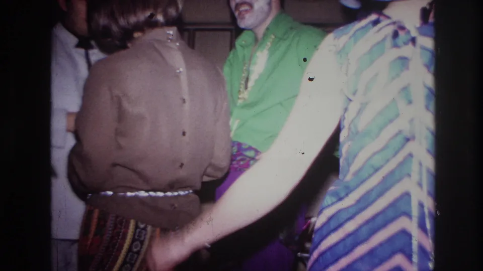 Ottawa 1967 home-movie frame of party guests standing close together, with a man in a green shirt and a striped dress passing near the camera.