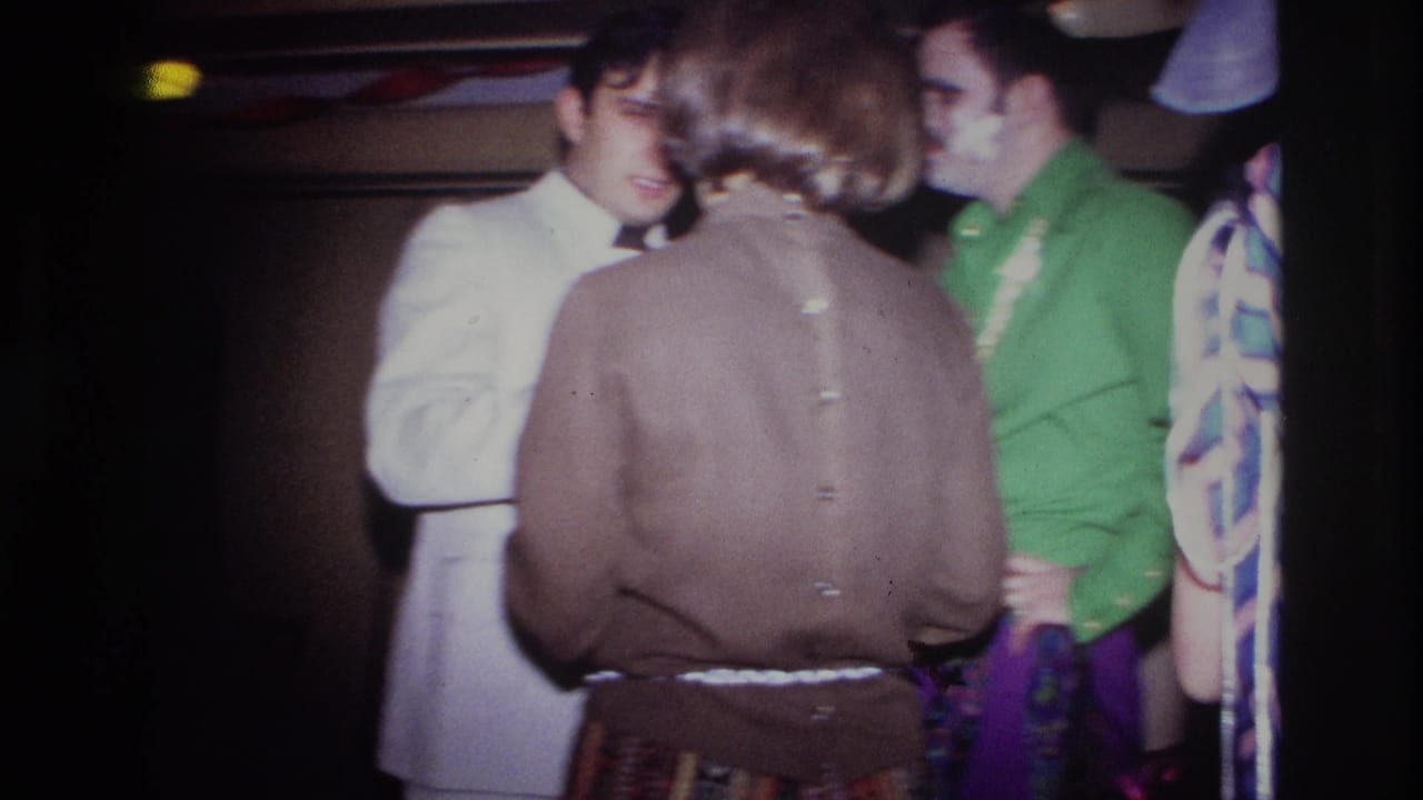 Party guests stand close together, including a man in a white suit and a man in a green shirt.