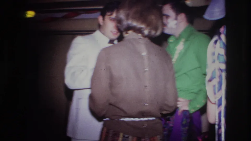 1967 Ottawa indoor birthday party scene with adults in period clothing, including a white suit and green shirt, seen in soft handheld footage.