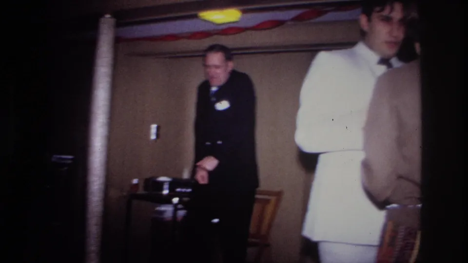 Home-movie view of an Ottawa party interior in 1967, with a dark-suited man near chairs and a white-suited guest at right.
