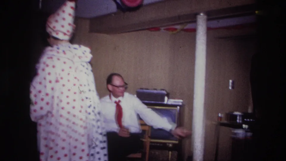 1967 Ottawa home-movie frame with a polka-dot clown costume, a seated adult man, chairs, wall paneling, and a television in the party room.