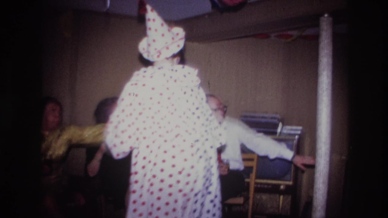 Clown in a white polka-dot costume stands before seated party guests indoors.