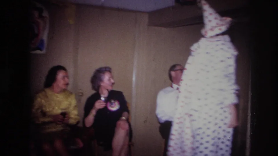 Adults in party clothing sit along a wood-paneled wall while a clown in a white polka-dot costume moves at the edge of the frame.