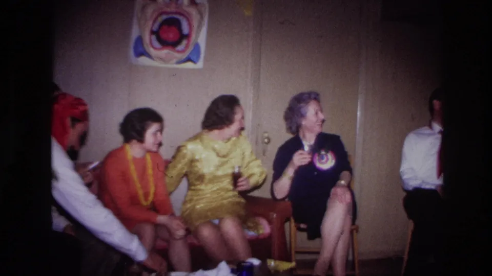 A group of adults in 1960s party clothing sit indoors below a clown-face decoration, with drinks visible and one person smoking.