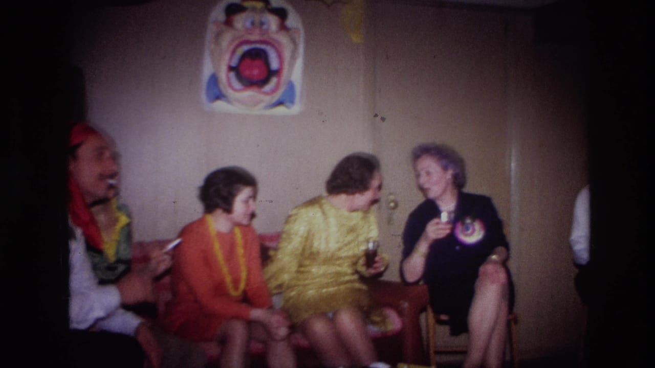 1967 Ottawa home-movie frame of adults in colorful party clothing seated in a wood-paneled basement with drinks and a wall poster.