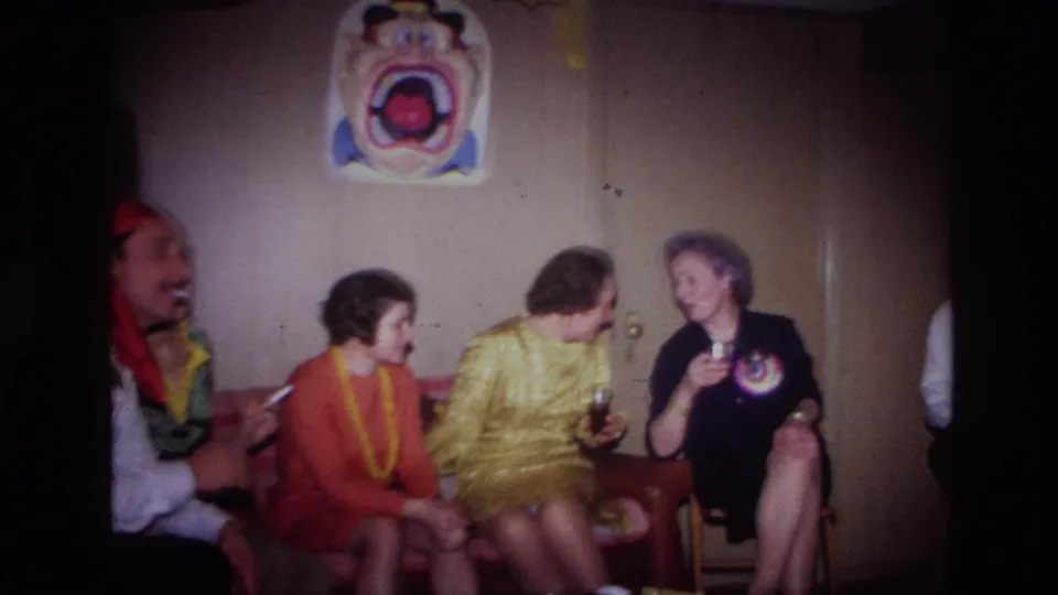 1967 Ottawa home-movie frame of adults in colorful party clothing seated in a wood-paneled basement with drinks and a wall poster.
