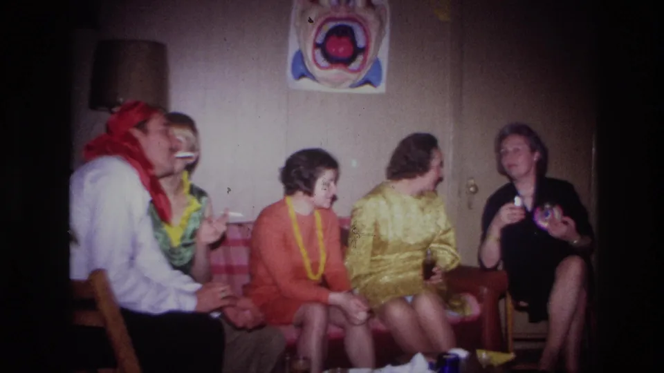 1967 Ottawa home-movie still showing adults seated in a basement party room, one holding a drink and another appearing to smoke.