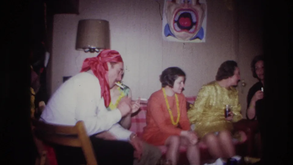 1967 Ottawa party footage frame with a man in a red bandana leaning toward seated women in a private basement room.