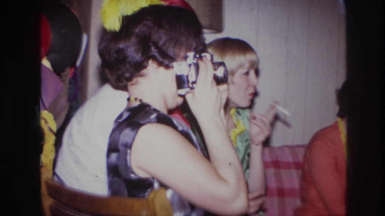 At a 1967 Ottawa indoor party, a woman in a sleeveless dress raises a compact camera as other guests sit nearby.