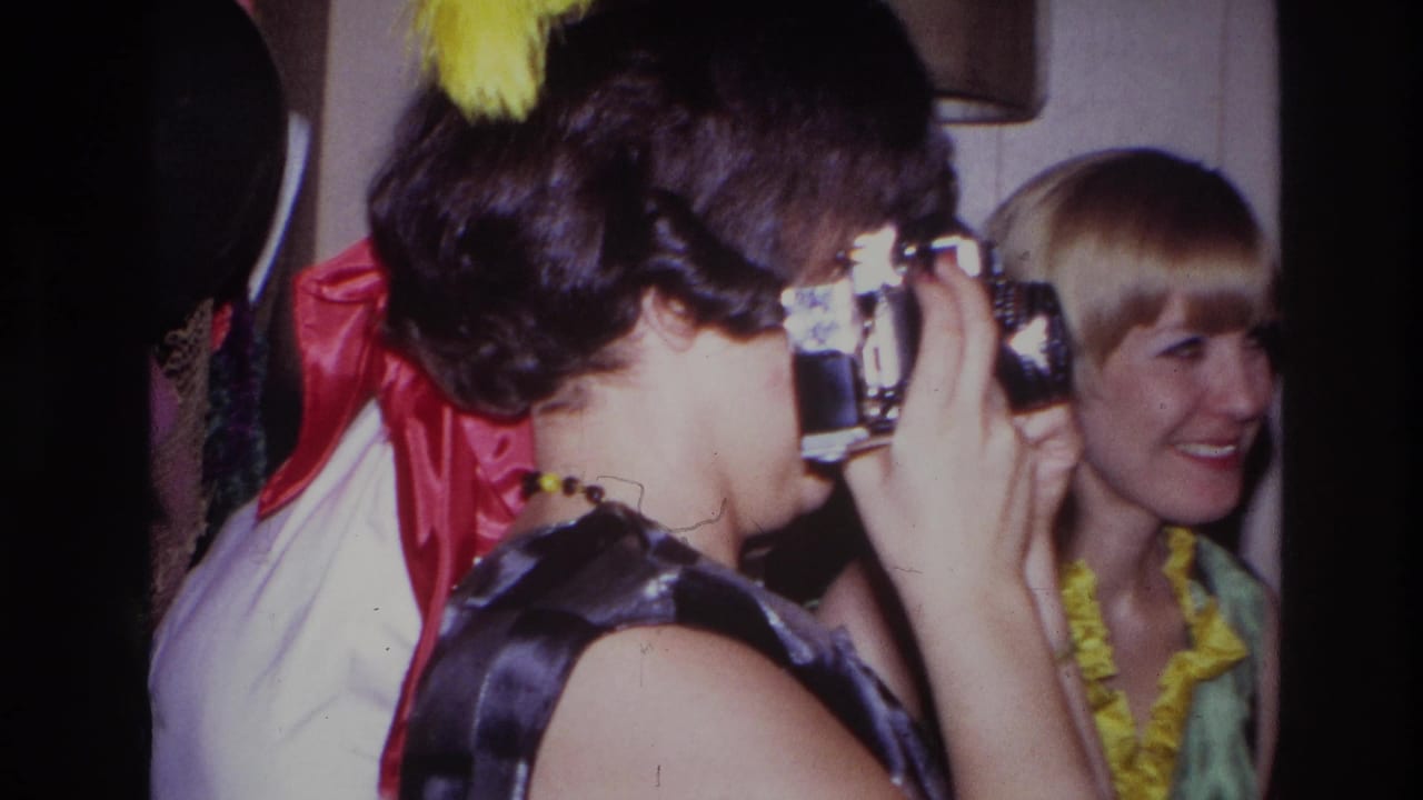 In a 1967 Ottawa home-movie party scene, a woman in a dark dress raises a compact camera while a blonde guest smiles beside balloons and decorations.