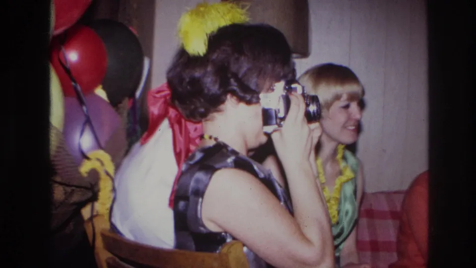 The woman continues aiming her camera during the Ottawa party, with a smiling blonde guest, balloons, and indoor decorations visible behind her.
