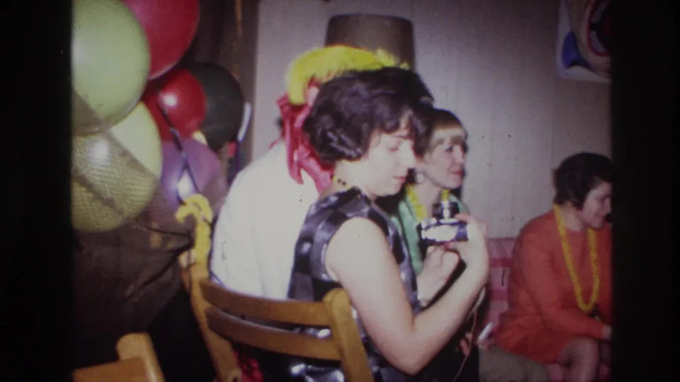 At the 1967 Ottawa party, the woman lowers her camera as other seated adults, balloons, leis, and costume accessories fill the private room.