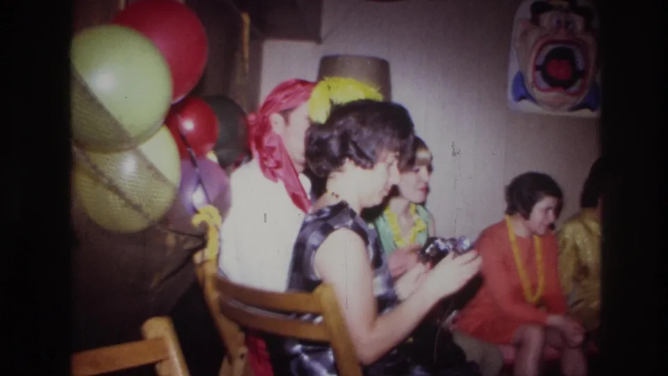 The woman looks down at the camera in her hands during the Ottawa home-movie party, with guests, balloons, and a clown face decoration nearby.