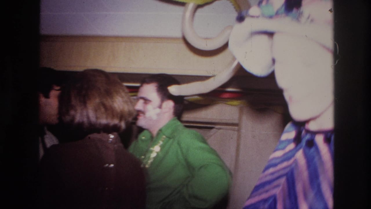 1967 Ottawa home-movie frame showing indoor party guests near streamers, with a smiling man in green and a woman turned away.