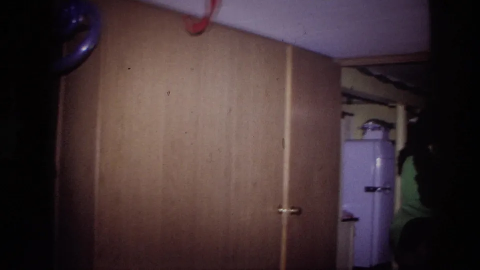 1967 Ottawa home-movie frame showing a dim wood-paneled party room with a white refrigerator and partial seated figure at the right edge.