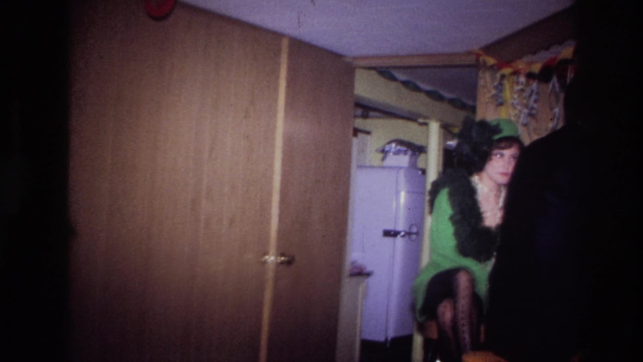 Woman in a green dress and feathered hat seated near a white refrigerator.