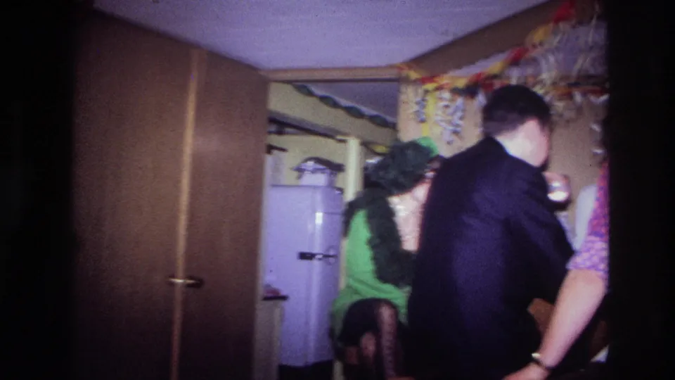 1967 Ottawa home-movie frame showing adults in party attire clustered in a wood-paneled room with bunting and a refrigerator behind them.