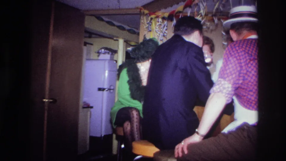 1967 Ottawa home-movie frame with adults in hats and dress clothes gathered closely in a private wood-paneled party room.