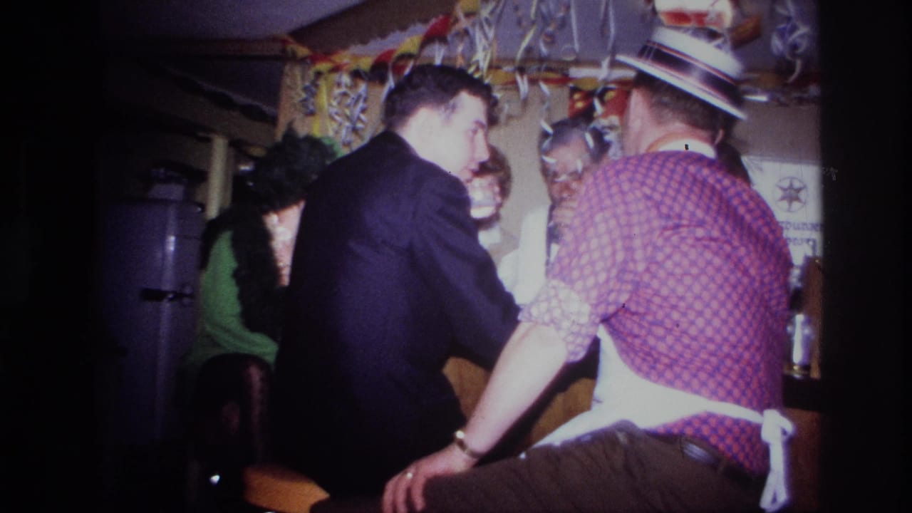 A man in a dark suit leans near a bar while another man in a checkered shirt sits beside him.