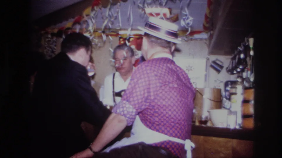 1967 Ottawa home-movie frame of a crowded bar scene with men in party attire, streamers, shelves, and cups visible.