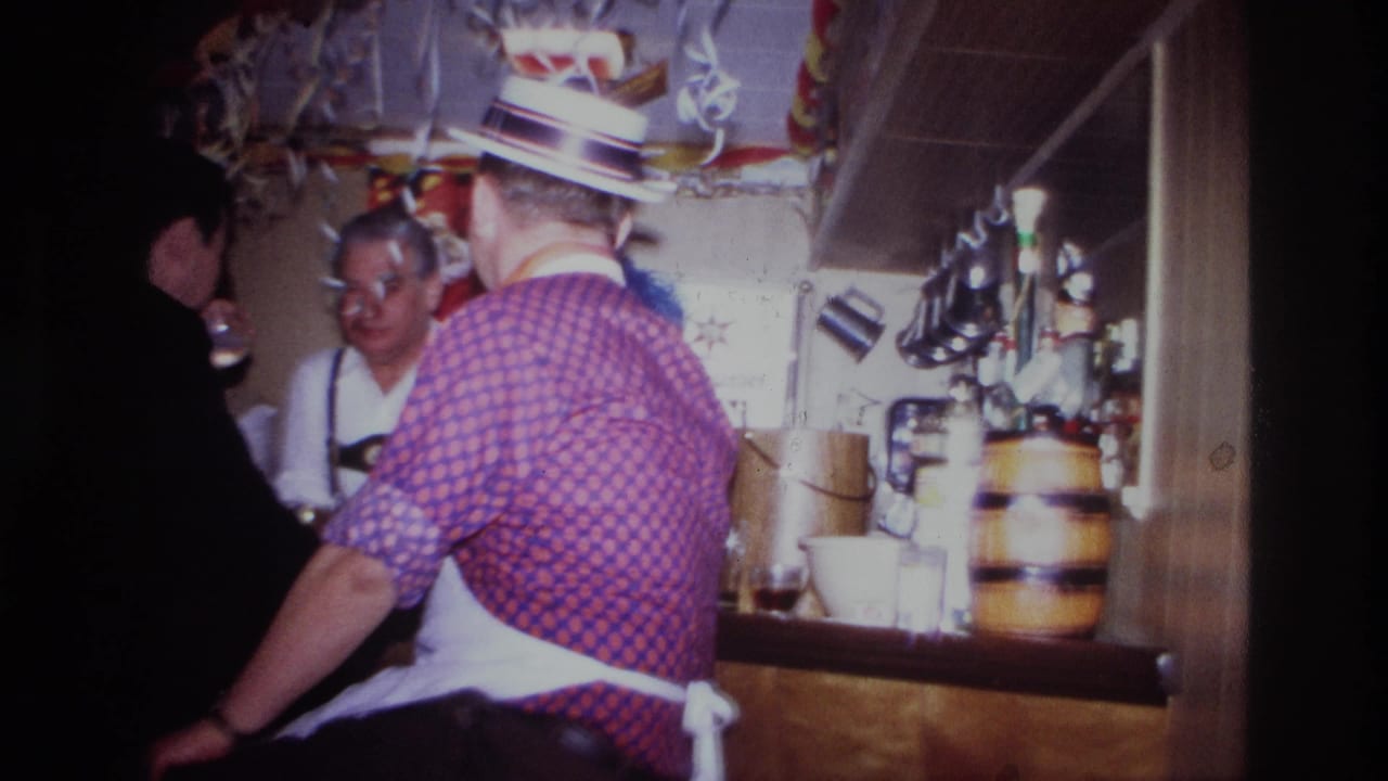 Men stand close together at a wooden bar beneath streamers and hanging barware.