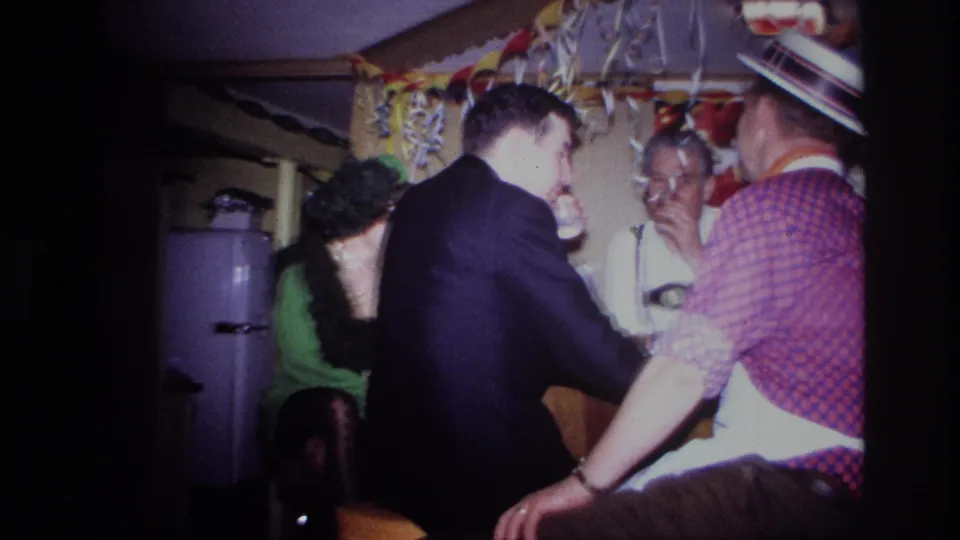 1967 Ottawa home-movie frame with adults gathered at a small bar interior, including a seated woman, refrigerator, streamers, and drinks.