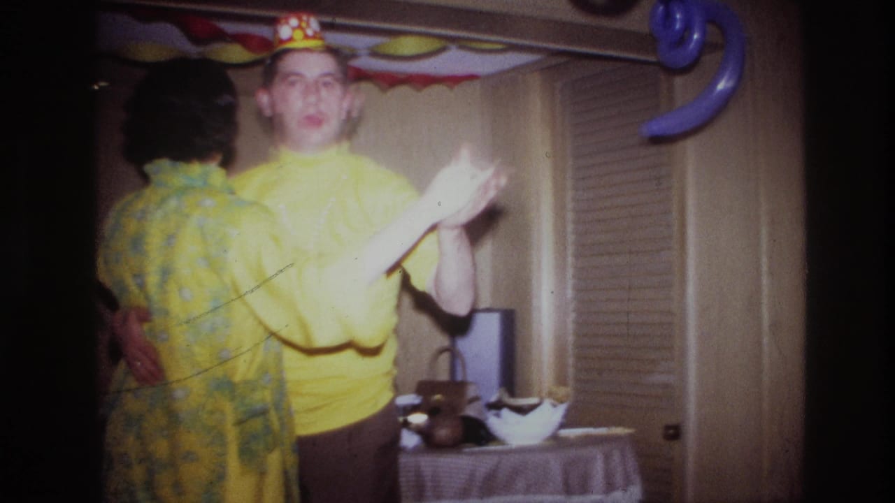 1967 Ottawa home-movie frame showing a couple dancing indoors, one in a yellow shirt and paper crown, with balloons and streamers behind them.