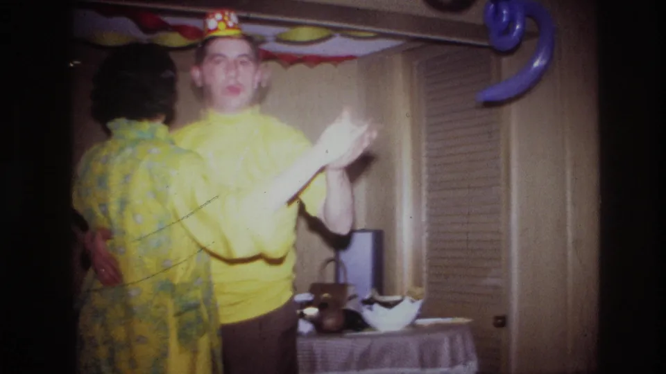 1967 Ottawa home-movie frame showing a couple dancing indoors, one in a yellow shirt and paper crown, with balloons and streamers behind them.