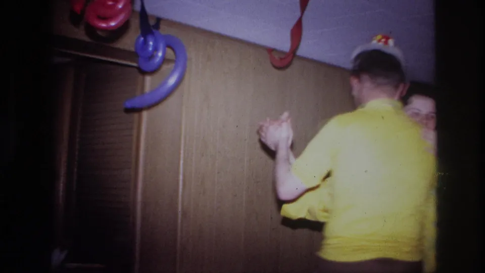 Two adults dance together in a 1967 Ottawa home-movie party scene with wood paneling, garlands, and hanging balloons.