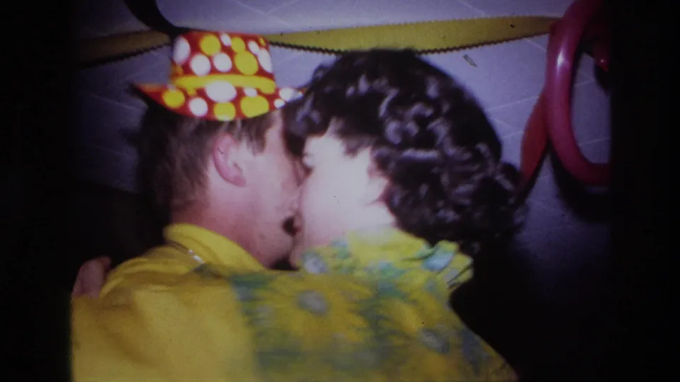 The young couple appears to kiss or embrace while dancing, with the party hat, yellow dress, and balloon decorations in view.