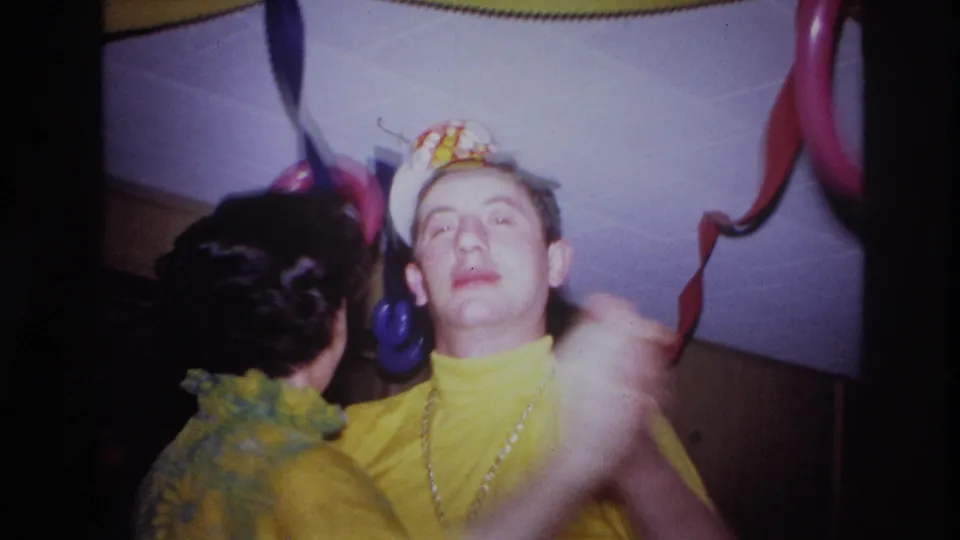 In this 1967 Ottawa home-movie frame, two adults in yellow party outfits dance closely beneath streamers and balloon decorations.
